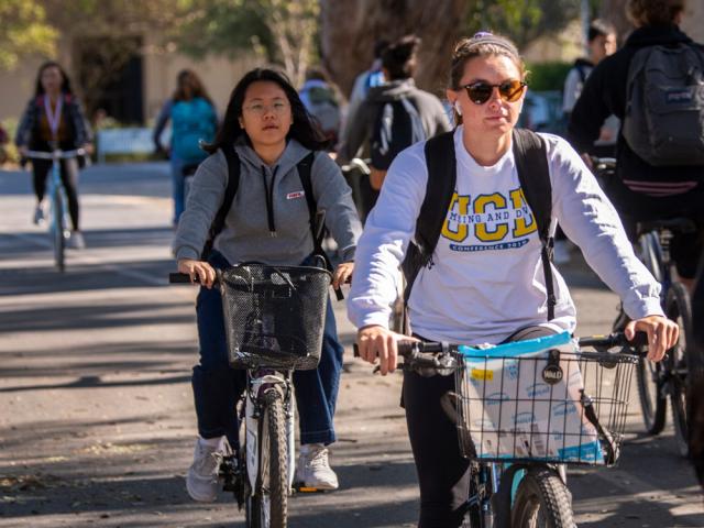 a closeup on two students riding among other students down a road on campus.