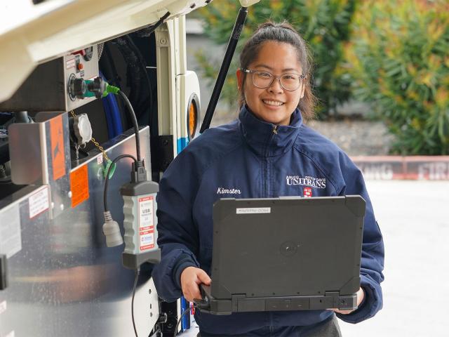A woman holds a computer outside a bus's rear panel that is open
