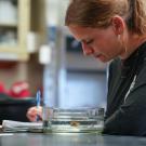Inside a laboratory setting, Zoe Brumbaugh watches an anemone in a dish next to her and takes notes with a pen and paper. 