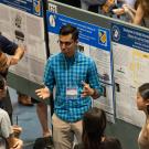 Student in blue short explains his research to a half-circle of people gathered round poster display